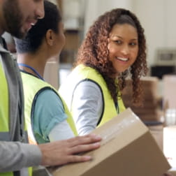 one young black man and two young black women working at a packaging facility. each is wearing a yellow safety vest.